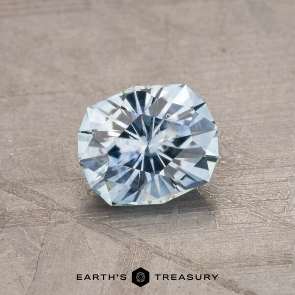 1.02-Carat Montana Sapphire (Heated) A 1.79-carat teal blue Montana sapphire (heated) is displayed against a textured gray background. The faceted gemstone, with its radiant cut, features intricate facets that catch and reflect light beautifully. The text "EARTH'S TREASURY" is written at the bottom in black.