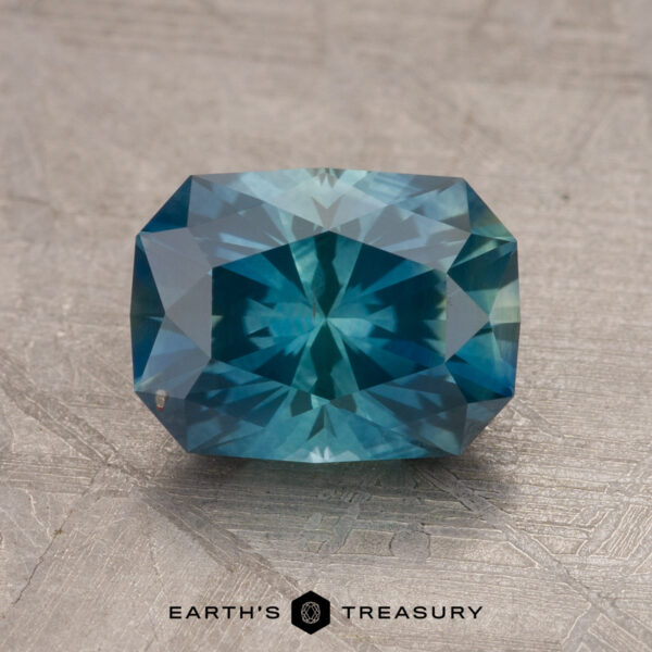 2.25-Carat Montana Sapphire (Heated) A 1.79-carat teal blue Montana sapphire, featuring a rectangular cut with facets that reflect various shades of blue, is set against a textured beige background. At the bottom, the text reads "EARTH'S TREASURY" alongside a small geometric logo.