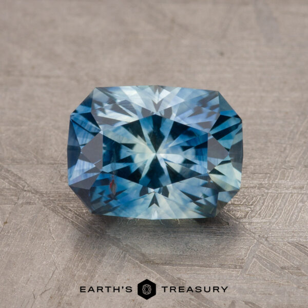 2.20-Carat Montana Sapphire (Heated) A stunning 1.79-carat teal blue Montana sapphire with a radiant star-like pattern is showcased on a textured surface against a neutral background. The text at the bottom reads "EARTH'S TREASURY".