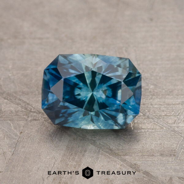 1.42-Carat Montana Sapphire (Heated) A vibrant 1.79-carat teal blue Montana sapphire (heated) with a rectangular shape and intricate internal facets is displayed on a textured gray surface. The gemstone exhibits a blend of deep and light blue hues, and "Earth's Treasury" is written at the bottom of the image.