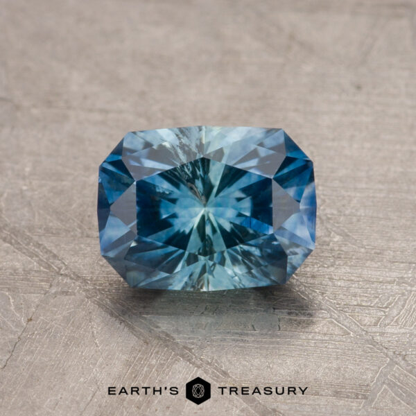 1.39-Carat Montana Sapphire (Heated) A dazzling, rectangular cushion-cut 1.79-carat Teal Blue Montana Sapphire (Heated) with intricate facets, displayed on a textured gray surface. The gem showcases a vibrant teal blue color with a radiant internal sparkle. Below the gem, the text "EARTH'S TREASURY" is visible.