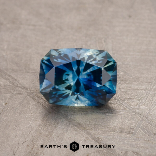 1.14-Carat Montana Sapphire (Heated) A 1.79-carat teal blue Montana sapphire, featuring a radiant cut style, is displayed against a textured background. The gemstone showcases deep blue and green hues with geometric facets. The text "EARTH'S TREASURY" is visible along the bottom edge of the image.