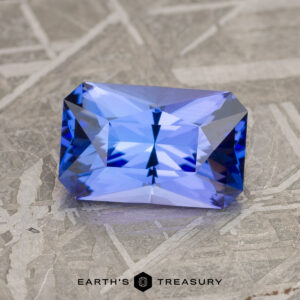 A 2.08-carat medium blue tanzanite with intricate facets, set against a textured gray background. The text "Earth's Treasury" appears at the bottom.