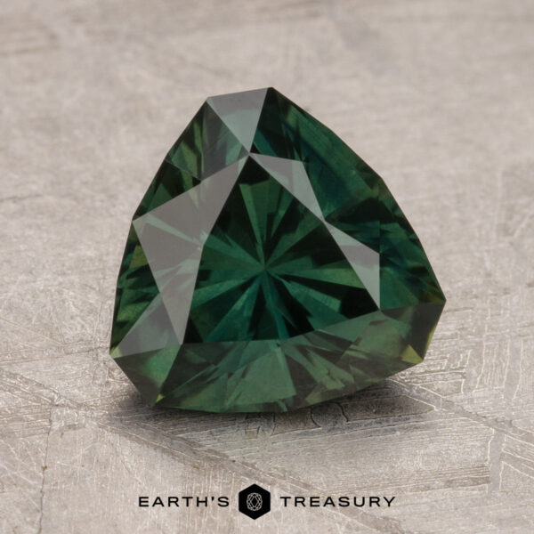 2.37-Carat Deep Bluish Green Australian Sapphire (Heated) A faceted, dark green 2.37-Carat Australian Sapphire in a triangular cut is displayed on a textured surface with the logo and text "Earth's Treasury" visible below it.
