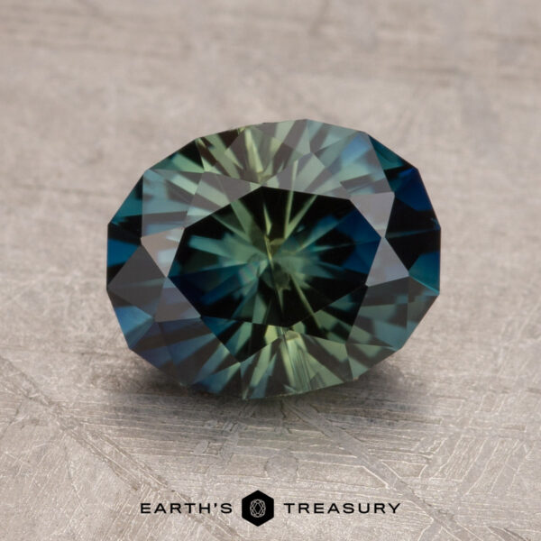 2.53-Carat Deep Blue-Green Particolored Australian Sapphire (Heated) A 2.53-carat Australian sapphire with a faceted oval shape and a blend of green and blue hues is placed on a textured surface. The words "Earth's Treasury" appear below the gemstone.