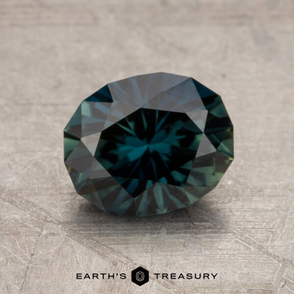 1.92-Carat Midnight Blue-Green Australian Sapphire (Heated) A 1.92-carat Australian sapphire with intricate cuts and a stunning blue-green hue, showcased on a textured gray surface. The gem beautifully reflects light, with the "Earth's Treasury" logo visible at the bottom.