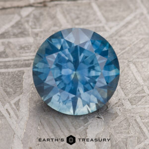 A 2.66-carat medium blue Montana sapphire (heated) is placed on a textured gray surface. Below the gemstone, the text "Earth's Treasury" is visible.