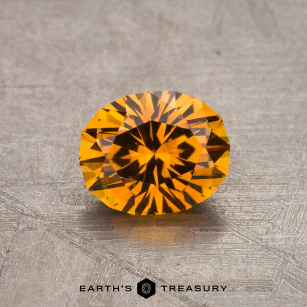 1.05-Carat Montana Sapphire (Heated) A round, faceted, 1.05-carat Orange Montana Sapphire (heated) with a vibrant, fiery color placed on a textured surface. The background is a grayish metallic material. Text at the bottom reads "Earth's Treasury" with a logo.