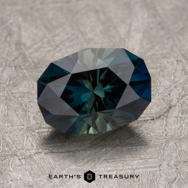 2.42-Carat Deep Blue-Olive Green Particolored Australian Sapphire (Heated) A 2.42-carat Australian sapphire is displayed on a textured surface, with the "Earth's Treasury" logo visible at the bottom center of the image.