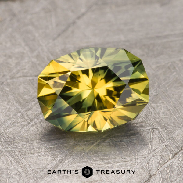 2.28-Carat Yellow-Green Particolored Australian Sapphire (Heated) A 2.28-carat Australian sapphire, featuring a faceted yellow-green hue with intricate cuts, rests on a textured metallic surface. The text "Earth's Treasury" is visible below the gemstone.