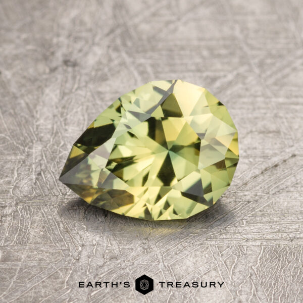 1.40-Carat Yellow-Green Particolored Australian Sapphire (Heated) A 1.40-carat Australian Sapphire, cut in a pear shape and faceted in a brilliant green hue, is displayed on a textured, gray surface. The text "EARTH'S TREASURY" appears at the bottom center of the image.