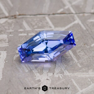 A 1.23-carat light violet-blue tanzanite gemstone with a hexagonal facet rests on a textured, light-colored surface. "EARTH'S TREASURY" is printed at the bottom of the image.