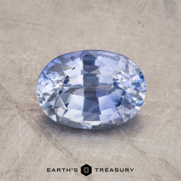 1.98-Carat Pale Blue Ceylon Sapphire (Heated) An oval-shaped faceted gemstone with a light blue color, identified as product JM1024L, displayed on a textured surface. The text "EARTH'S TREASURY" is visible at the bottom of the image.