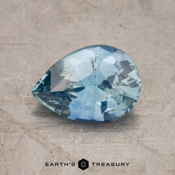 1.59-Carat Aqua Blue Sapphire (Heated) A pear-shaped blue gemstone (Product Code: JM1028L) rests on a textured surface. The image includes the logo and the text "Earth's Treasury" at the bottom.