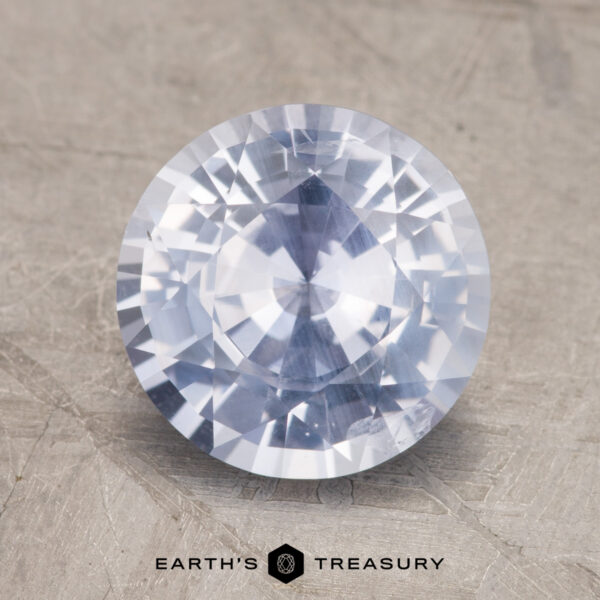 3.06-Carat Near Colorless Sapphire A close-up image of a faceted 3.06-Carat Sapphire, its blue hue glistening on a textured surface. At the bottom, the text reads "EARTH'S TREASURY.