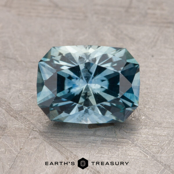 2.14-Carat Robin’s Egg Blue Montana Sapphire (Heated) A 2.14-carat Montana Sapphire (heated), featuring a rectangular, faceted blue gemstone, is placed on a textured surface. The bottom center displays the logo "Earth's Treasury.
