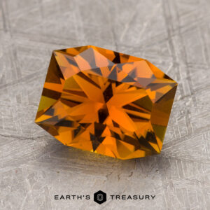 A faceted orange 2.37-carat tourmaline lies on a textured gray surface. The bottom part of the image features the text "Earth's Treasury.