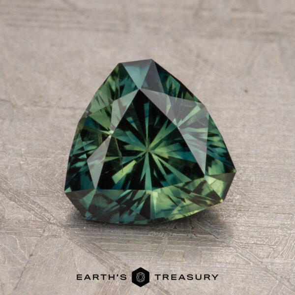 1.98-Carat Australian Sapphire (Heated) A close-up view of a polished 1.98-Carat Australian Sapphire (Heated) with a triangular shape on a textured gray surface. The text "Earth's Treasury" is visible at the bottom.