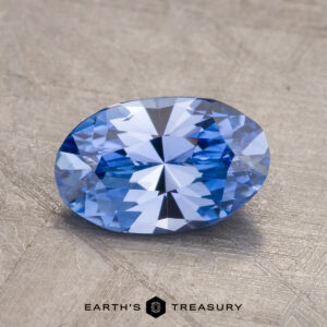 A 1.57-carat Ceylon Sapphire, expertly faceted into an oval shape, is displayed against a textured gray background with the "EARTH'S TREASURY" logo prominently positioned at the bottom center.