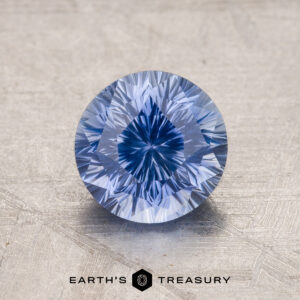 A close-up image of a round brilliant-cut blue gemstone on a textured surface with the text "1.99-Carat Ceylon Sapphire" at the bottom.