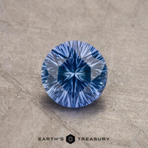 A brilliant 1.04-carat, round-cut Rich Sky Blue Ceylon Sapphire sits on a textured, lightly scuffed surface. The gemstone's facets create a striking, star-like pattern in its center. The words "EARTH'S TREASURY" are written below the gemstone.