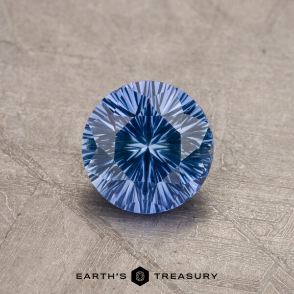1.04-Carat Ceylon Sapphire A brilliant 1.04-carat, round-cut Rich Sky Blue Ceylon Sapphire sits on a textured, lightly scuffed surface. The gemstone's facets create a striking, star-like pattern in its center. The words "EARTH'S TREASURY" are written below the gemstone.