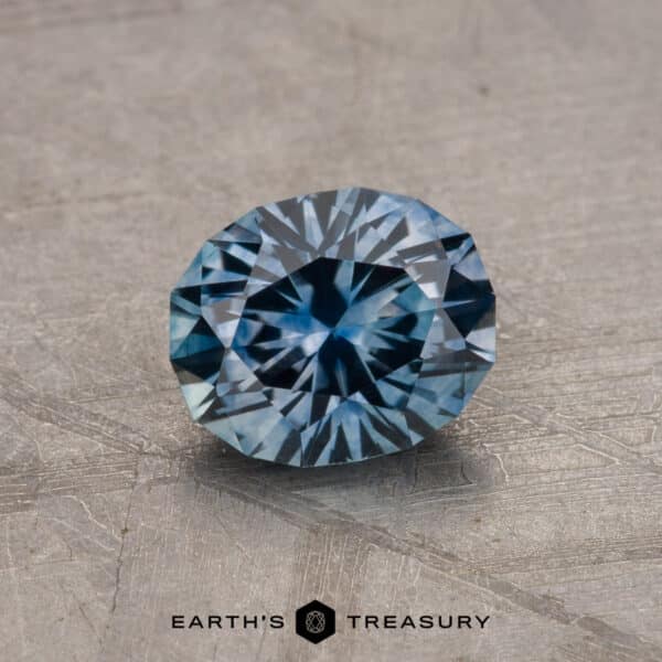 1.21-Carat Steely Blue Montana Sapphire (Heated) A round cut 1.21-carat Montana Sapphire (Heated) displayed on a textured surface with the text “EARTH’S TREASURY” at the bottom.