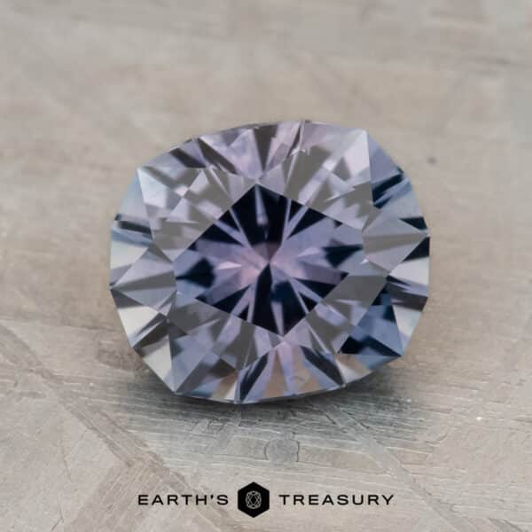 2.14-Carat Montana Sapphire (Heated) A faceted gemstone with a clear, multi-faceted surface rests on a textured gray background. The text "Earth's Treasury" is visible beneath the gemstone, which stunningly exhibits the steely blue to purple color change of a heated 2.14-carat Montana sapphire.