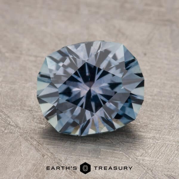 2.14-Carat Montana Sapphire (Heated) A round, steely blue to purple faceted sapphire is displayed on a textured surface. The text below reads "EARTH'S TREASURY 2.14-Carat Montana Sapphire (Heated).