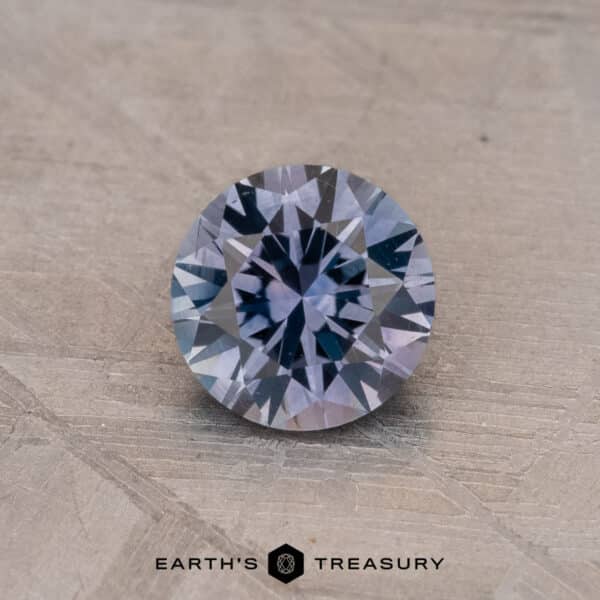 0.84-Carat Montana Sapphire (Heated) A 0.84-carat round-cut Montana Sapphire (Heated) with a violet hue sits on a textured surface. The words "Earth's Treasury" are visible at the bottom of the image.