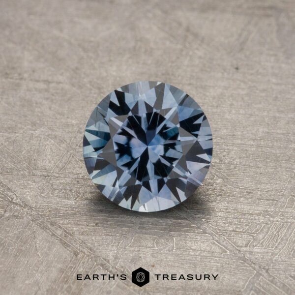 0.84-Carat Montana Sapphire (Heated) A 0.84-carat Montana Sapphire (Heated) with intricate facets is displayed on a textured surface, showcasing its steely blue hue. The text "EARTH'S TREASURY" is visible at the bottom.