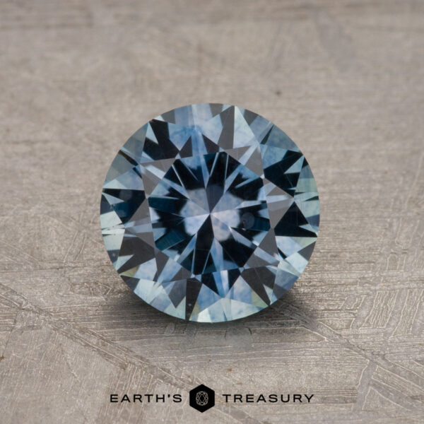 1.60-Carat Montana Sapphire (Heated) A round, faceted 1.60-carat Montana Sapphire (Heated) with shades of blue sits on a textured surface, with "Earth's Treasury" written at the bottom in black text.