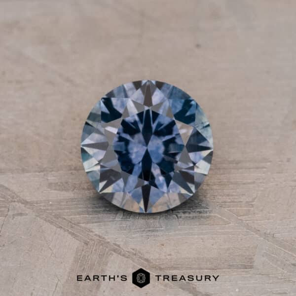0.92-Carat Montana Sapphire (Heated) A round faceted gemstone with a steely blue and gray hue, resting on a textured surface. The text "Earth's Treasury" is displayed below the 0.92-Carat Montana Sapphire (Heated).