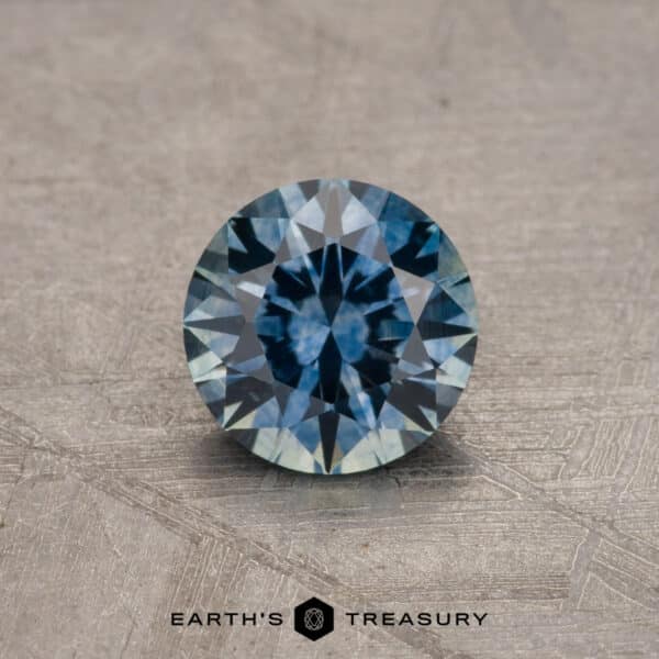 0.92-Carat Montana Sapphire (Heated) A round-cut blue-green Montana Sapphire gemstone, weighing 0.92 carats, is displayed on a textured surface. The bottom of the image features the logo and text "Earth's Treasury.