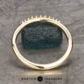 The "Paloma" band in 14k yellow gold with empty prongs stands upright, a raw blue gemstone behind it and "EARTH'S TREASURY" text at the bottom.