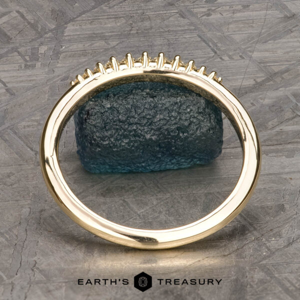 The "Paloma" band in 14k yellow gold The "Paloma" band in 14k yellow gold with empty prongs stands upright, a raw blue gemstone behind it and "EARTH'S TREASURY" text at the bottom.