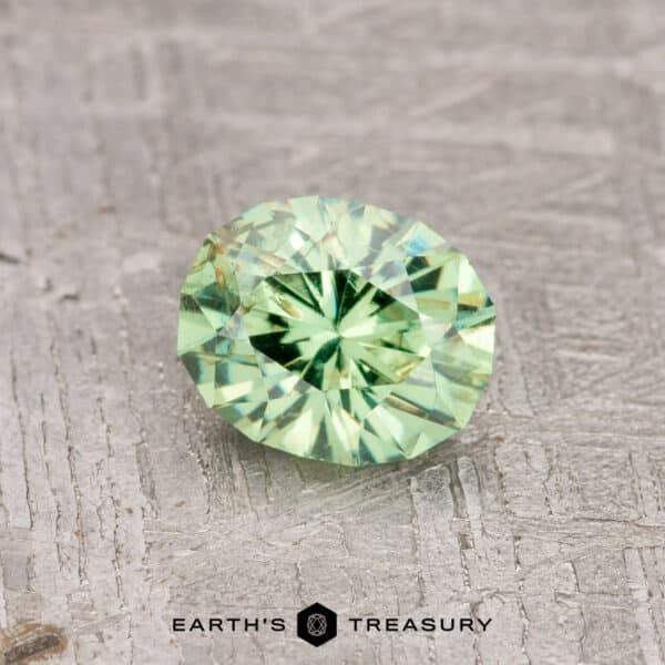 0.84-Carat Mint Green Demantoid Garnet A 0.84-carat mint green demantoid garnet with a round, faceted design is showcased on a textured gray surface. Below it, you'll find the Earth's Treasury logo.