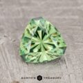 A faceted, triangular 1.57-carat medium green demantoid garnet rests on a textured gray surface. Text reads: "Earth's Treasury.
