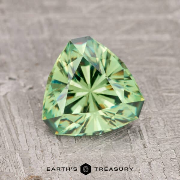 1.57-Carat Medium Green Demantoid Garnet A faceted, triangular 1.57-carat medium green demantoid garnet rests on a textured gray surface. Text reads: "Earth's Treasury.