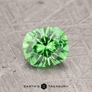 A faceted green gemstone on a textured surface, labeled "0.99-Carat Tsavorite Garnet.