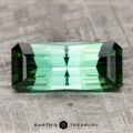 A rectangular 26.68-carat rich blue-green tourmaline with multiple facets rests on a textured, gray surface. The text "Earth's Treasury" is visible beneath the gemstone.