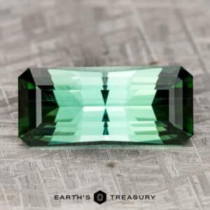 A rectangular 26.68-carat rich blue-green tourmaline with multiple facets rests on a textured, gray surface. The text "Earth's Treasury" is visible beneath the gemstone.