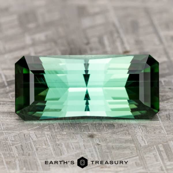 26.68-Carat Rich Blue-Green Tourmaline A rectangular 26.68-carat rich blue-green tourmaline with multiple facets rests on a textured, gray surface. The text "Earth's Treasury" is visible beneath the gemstone.