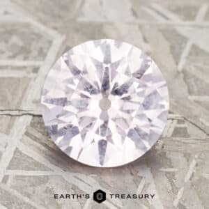 A 2.91-carat pale pink Montana sapphire, featuring a round faceted design and a light, translucent appearance, rests on a textured gray surface.