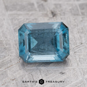 A rectangular, teal aqua gemstone with step-cut facets is placed on a textured gray surface. The text "1.59-Carat Nasarawa" appears below the gem.