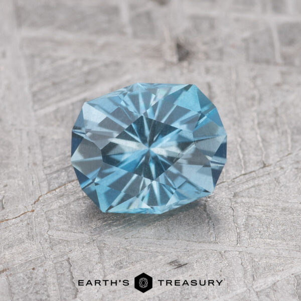 0.92-Carat Nasarawa Teal Aqua The 0.92-Carat Nasarawa Teal Aqua, a round, faceted blue gemstone, is showcased on a textured gray background with the Earth’s Treasury logo visible at the bottom.