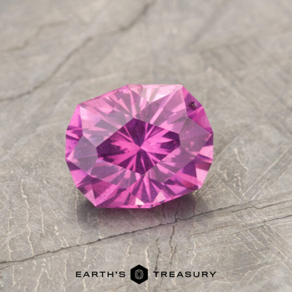 1.29-Carat Pastel Pyrope Color-Change Garnet Product ET1127, a faceted gemstone with a purple-pink hue and an intricate cut, is situated on a textured gray surface, showcasing the Earth's Treasury logo beneath it.