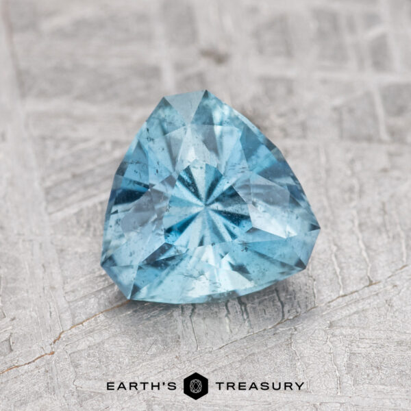 0.99-Carat Nasarawa Teal Aqua A trillion-cut 0.99-carat Nasarawa Teal Aqua gemstone on a textured gray surface, featuring the "Earth's Treasury" logo at the bottom.