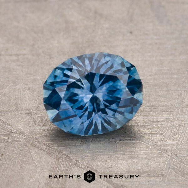 1.21-Carat Montana Sapphire (Heated) A round brilliant 1.21-carat Montana Sapphire gemstone, heated for enhanced quality, placed on a textured gray surface with the text "Earth's Treasury" beneath.