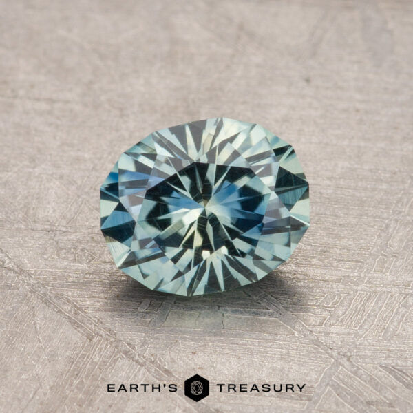 1.13-Carat Montana Sapphire (Heated) 1.13-carat oval-cut Montana sapphire featuring a bluish-green hue, placed on a textured surface with the label "Earth's Treasury" at the bottom.